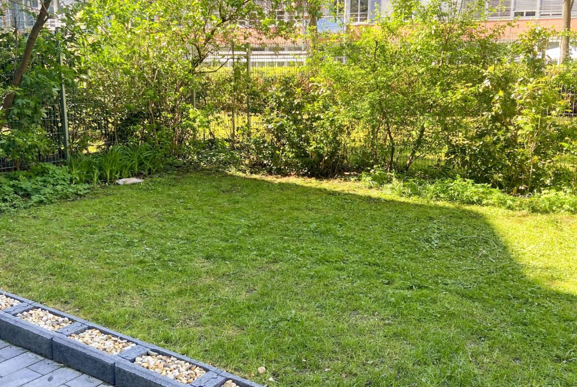 Green lawn with a dense hedge and metal fence behind, edge bordered by pebble-filled planters along a paved walkway.