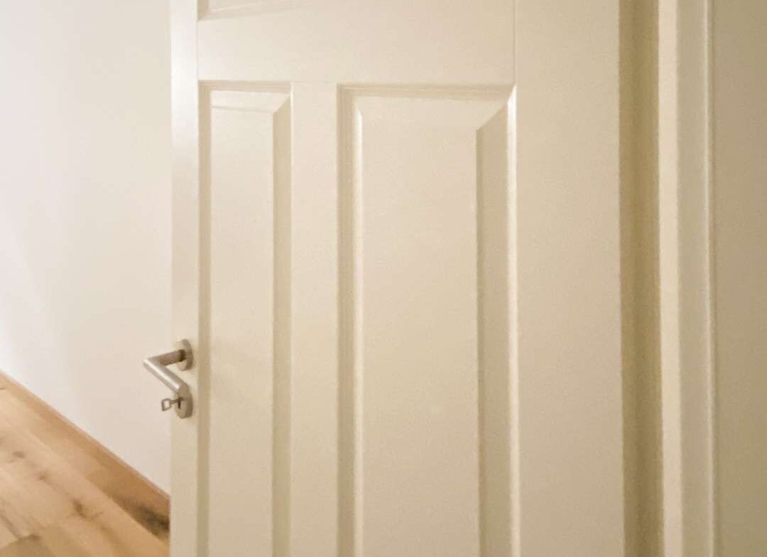 White paneled interior door partially open in a bright hallway with light wood flooring and a silver handle.