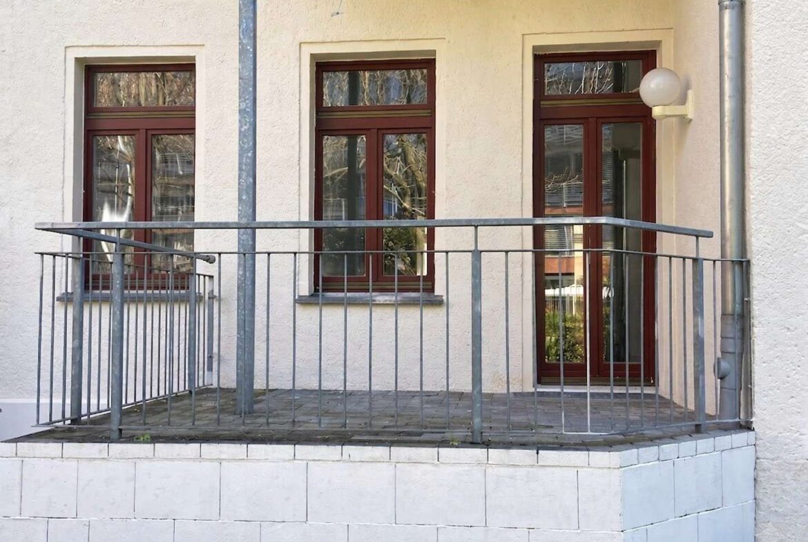 Small balcony with a metal railing in front of a beige stucco building, featuring two large windows and a glass door with brown frames.