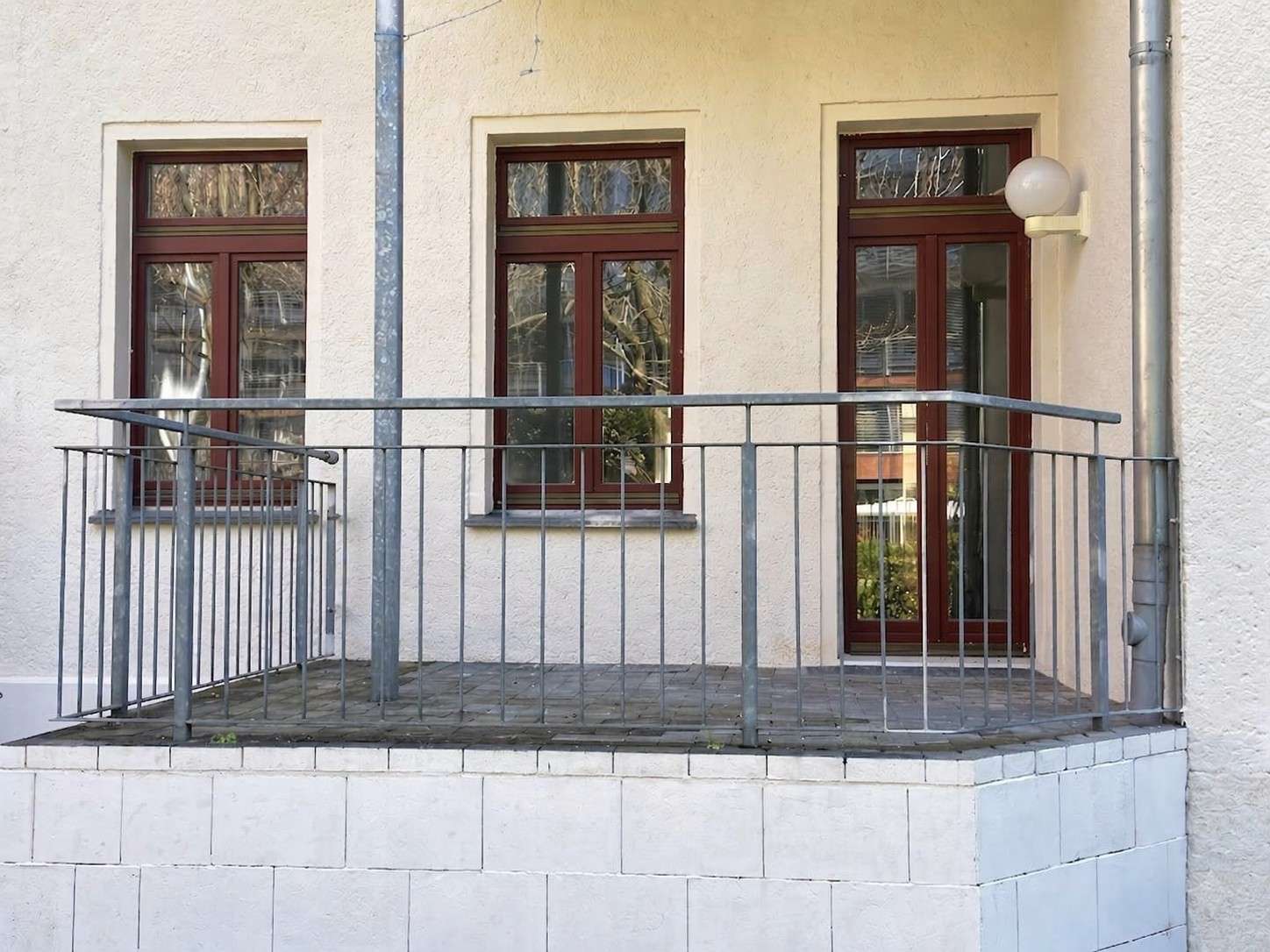 Small balcony with a metal railing in front of a beige stucco building, featuring two large windows and a glass door with brown frames.