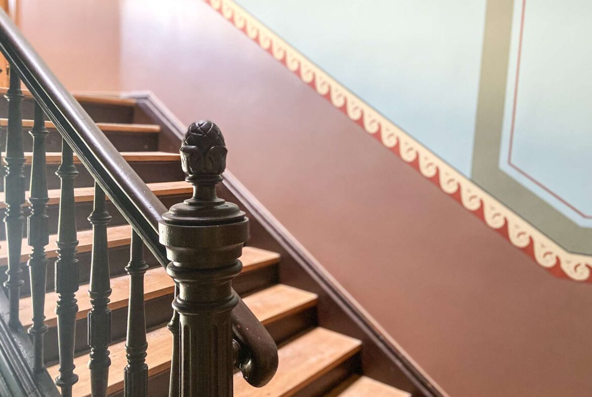 Close-up of a dark metal staircase railing with a carved newel post and wooden steps along a wall with patterned red trim.