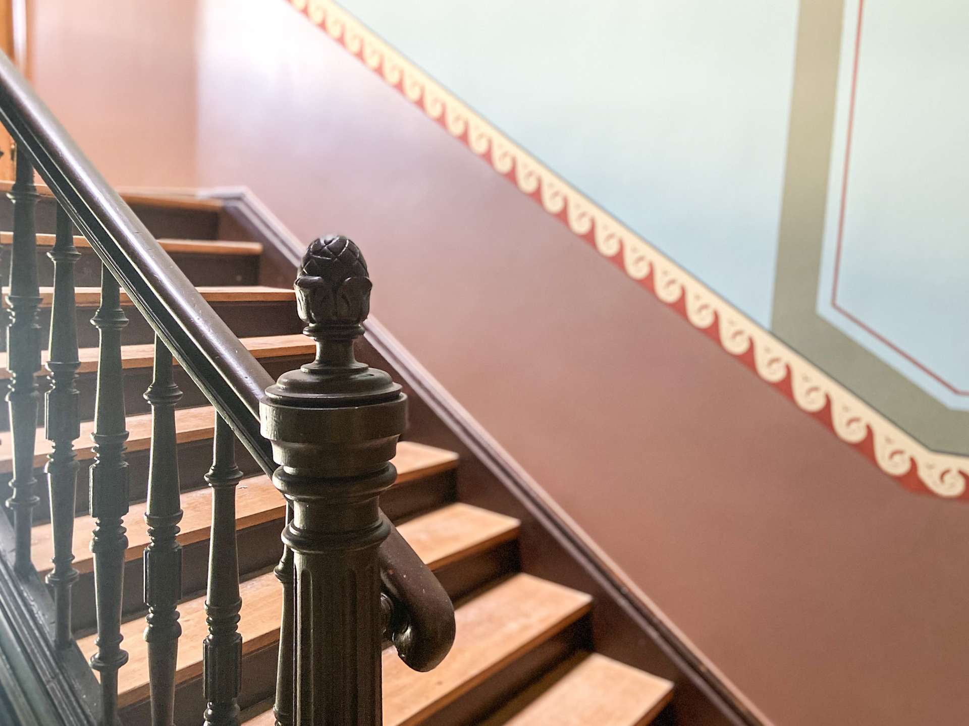Close-up of a dark metal staircase railing with a carved newel post and wooden steps along a wall with patterned red trim.