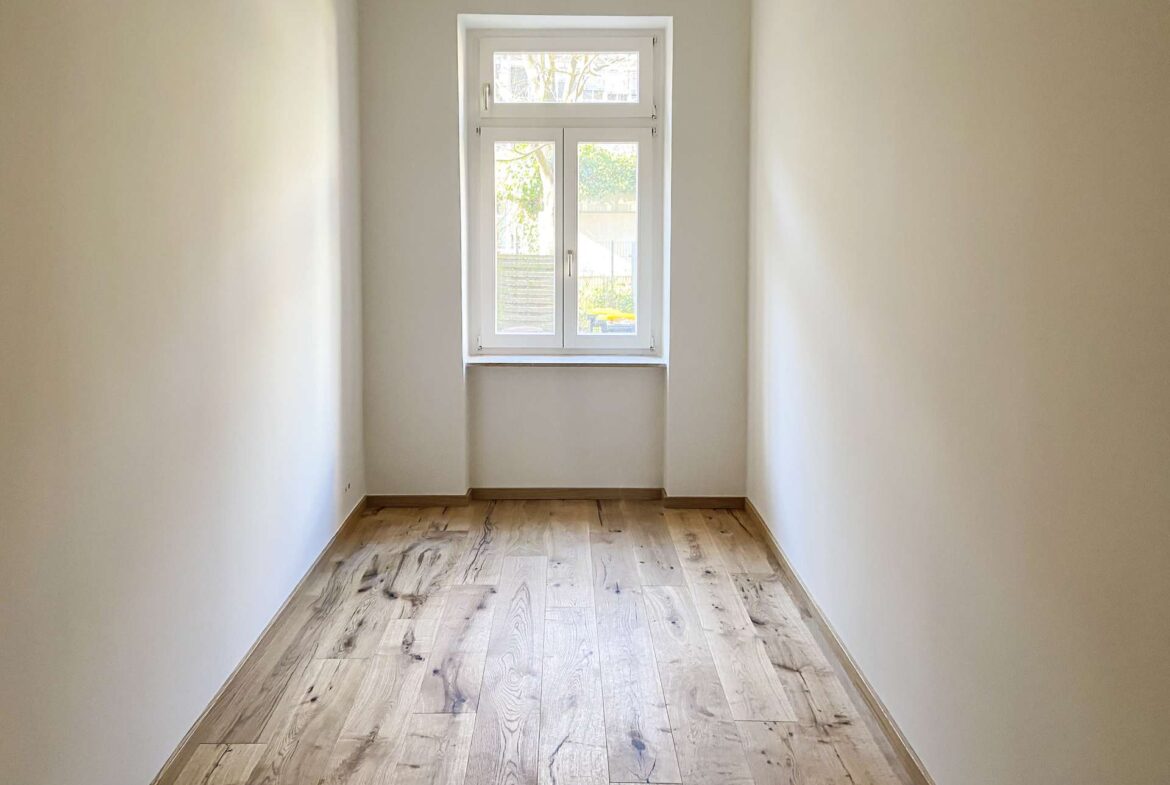 Narrow empty hallway with light wood flooring and a window at the far end.