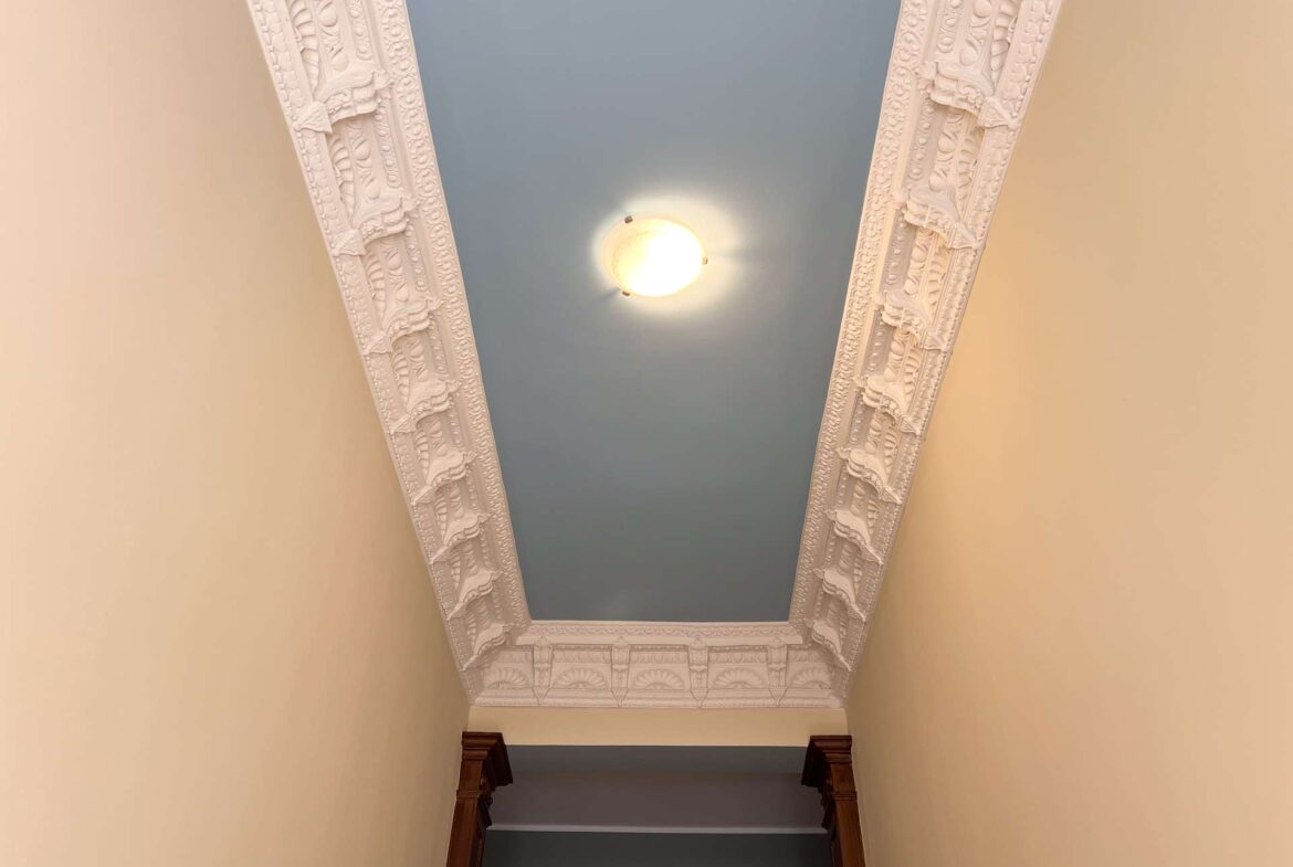 Ornate white plaster molding frames a blue ceiling in a narrow stairwell; beige walls and a round ceiling light.
