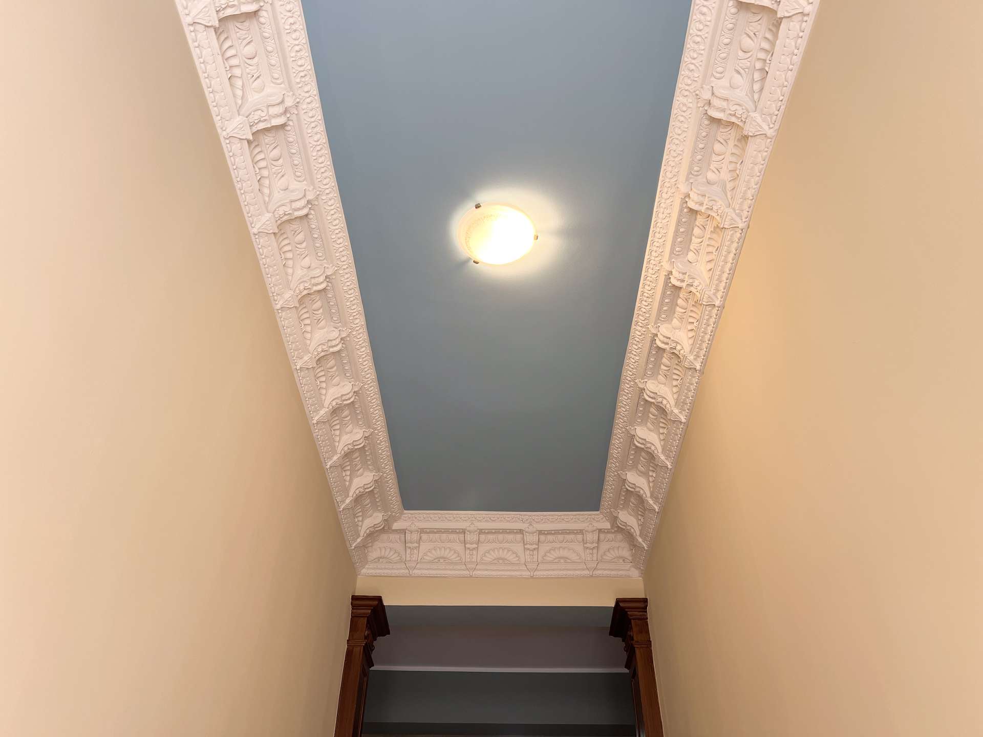 Ornate white plaster molding frames a blue ceiling in a narrow stairwell; beige walls and a round ceiling light.