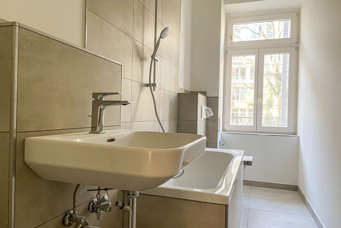 Modern bathroom featuring a wall-mounted rectangular sink with chrome faucet, exposed plumbing, tiled walls, and a large window bringing in daylight.