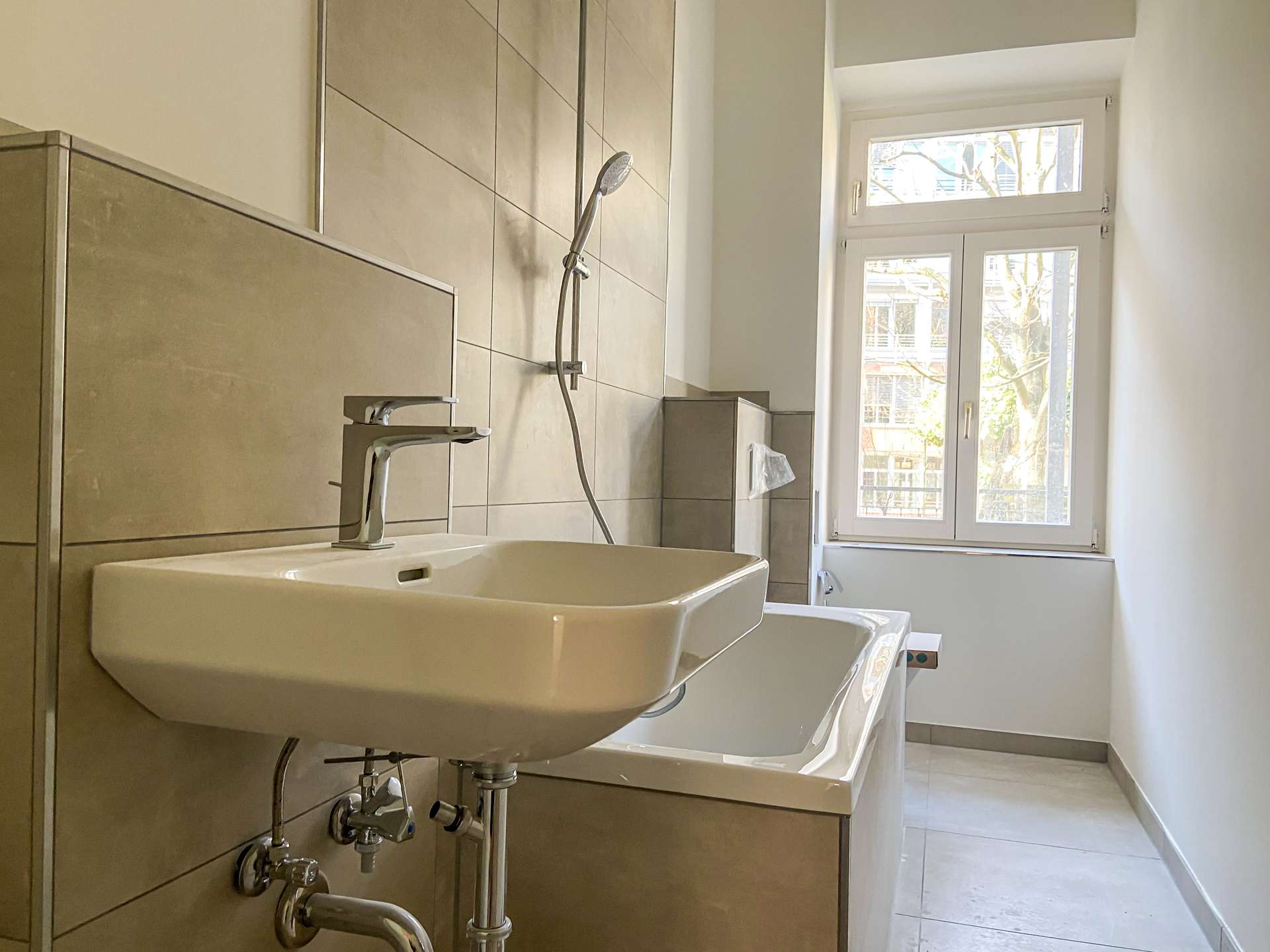 Modern bathroom featuring a wall-mounted rectangular sink with chrome faucet, exposed plumbing, tiled walls, and a large window bringing in daylight.