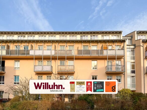 Apartment building with balconies and a large Willuhn Immobilien sign in front; real estate branding and awards displayed on the signboard