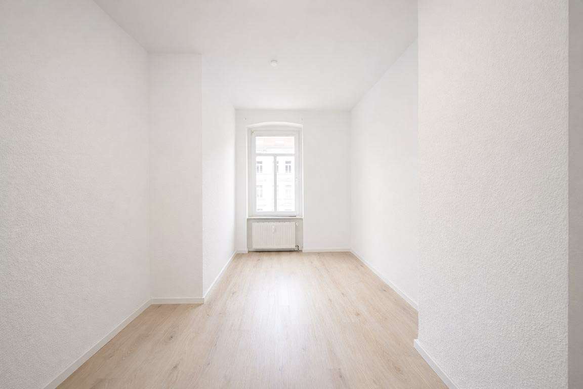 Empty narrow room with white walls, light wood flooring, and a window at the far end with a radiator underneath.