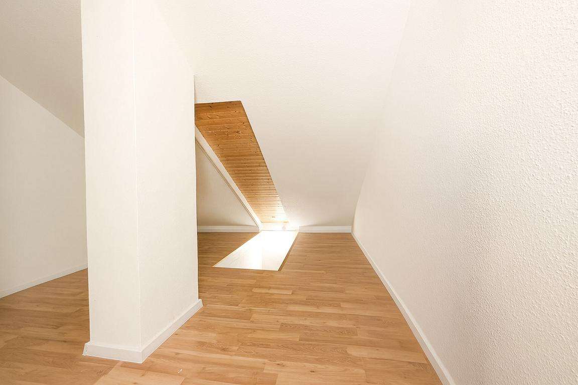 Attic room with a sloped ceiling, white walls, and light wooden flooring; sunlight filters through a skylight.
