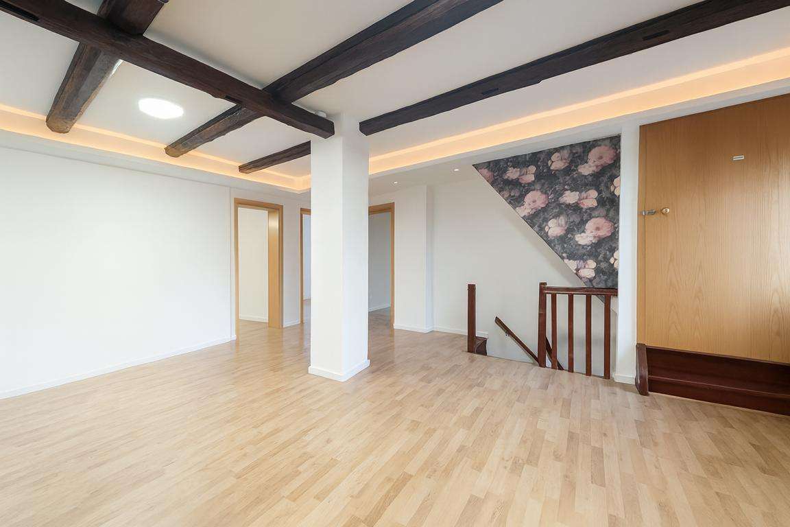 Spacious empty loft with exposed dark wooden beams, light wood flooring, white walls, and a staircase railing by a floral accent wall.