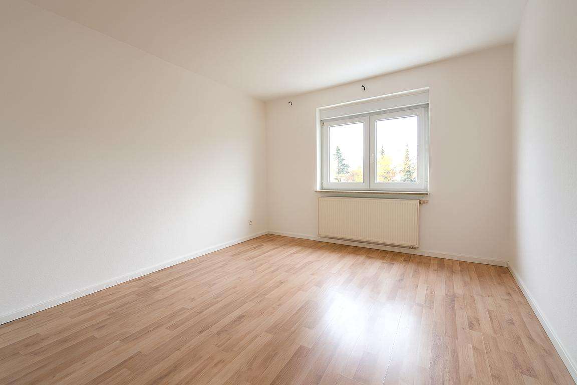 Bright, empty room with white walls, light wood flooring, and a double window with a radiator beneath it under soft natural light.