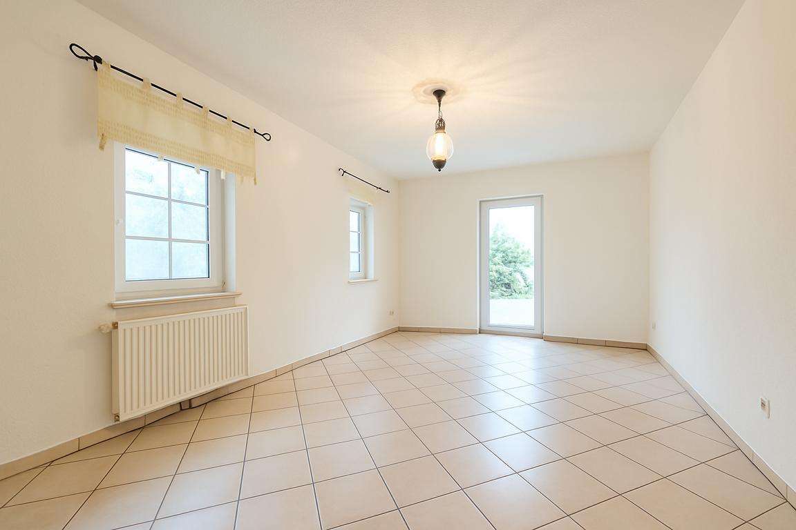 Bright empty living room with tiled floor, two windows, radiator, and a glass door to the outside.