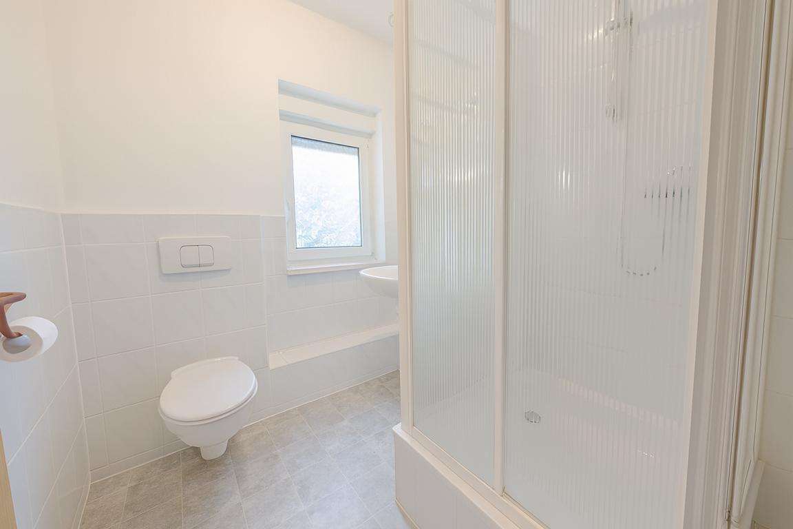 Bright white bathroom with a wall-mounted toilet, flush panel, a frosted-glass shower enclosure, and a small window on the wall.