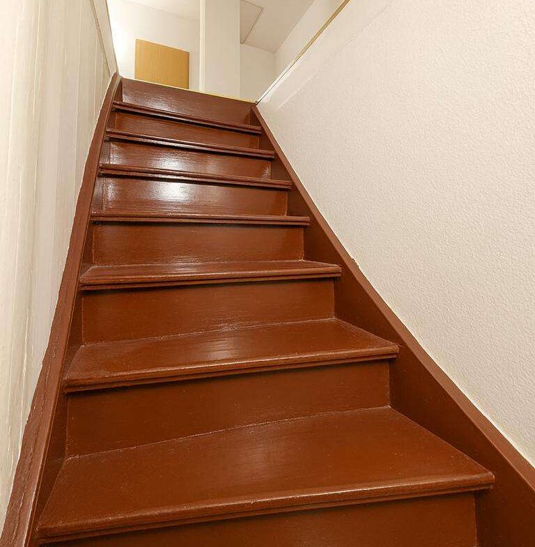 Brown wooden staircase ascending to a well-lit upper floor, with white walls on both sides.