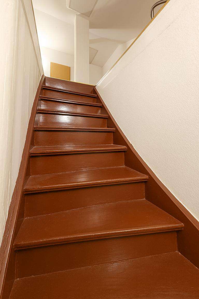 Brown wooden staircase ascending to a well-lit upper floor, with white walls on both sides.