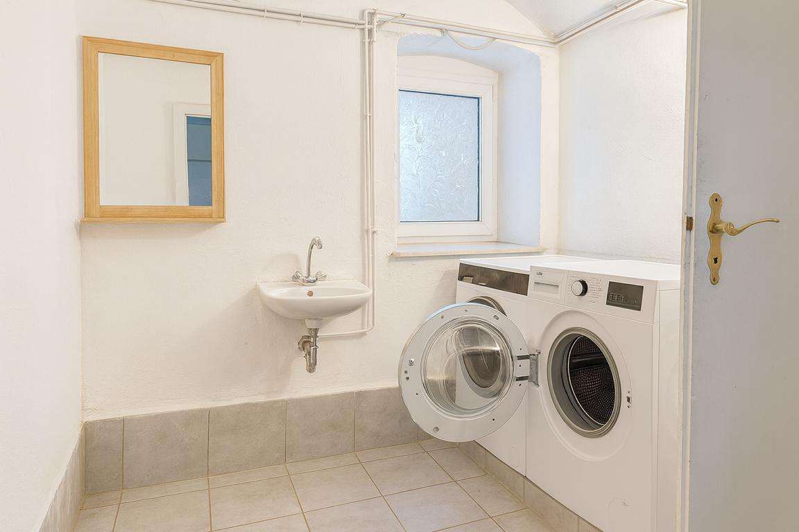 Laundry room with a front-loading washing machine, a small wall-mounted sink, a window, and a mirror on the wall.