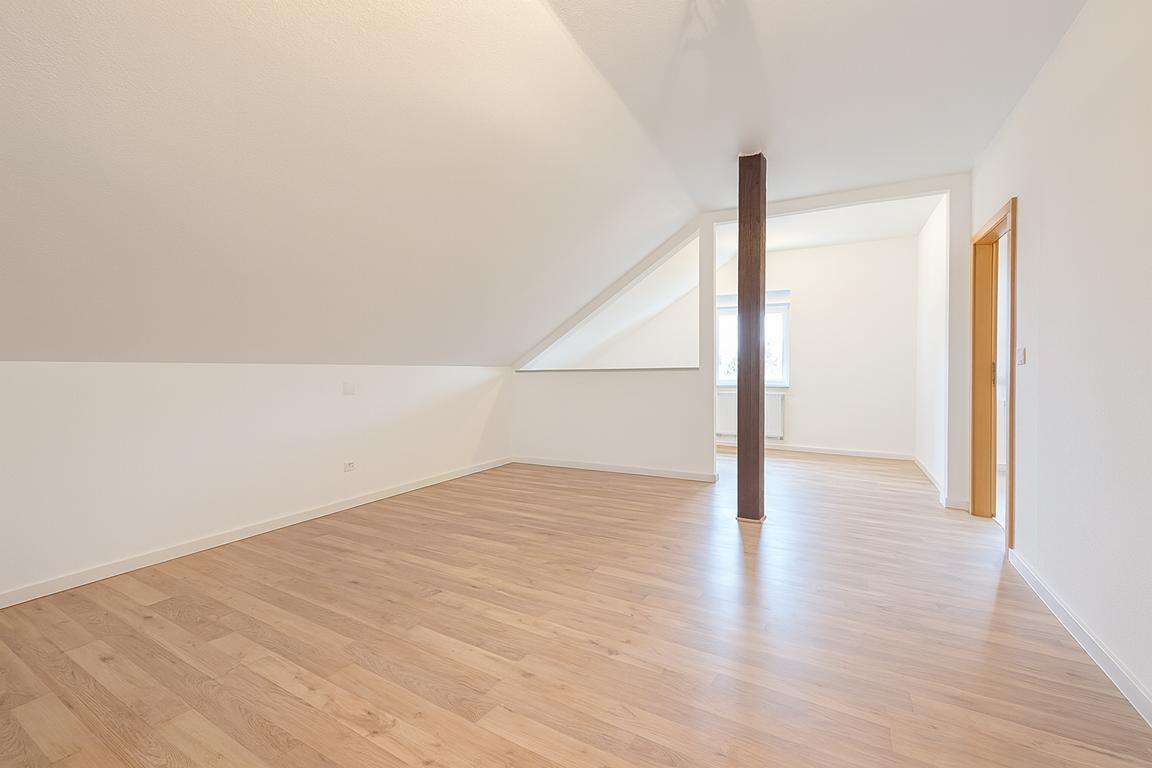 Bright empty attic room with a sloped ceiling, light wood flooring, and a dark central pillar; doorway leads to a sunlit adjacent room.