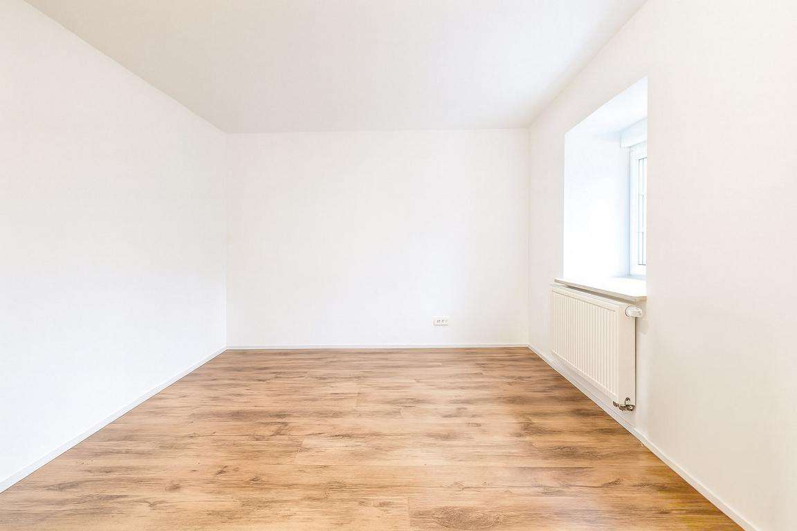 Empty white room with light wood flooring and a radiator under a window on the right.