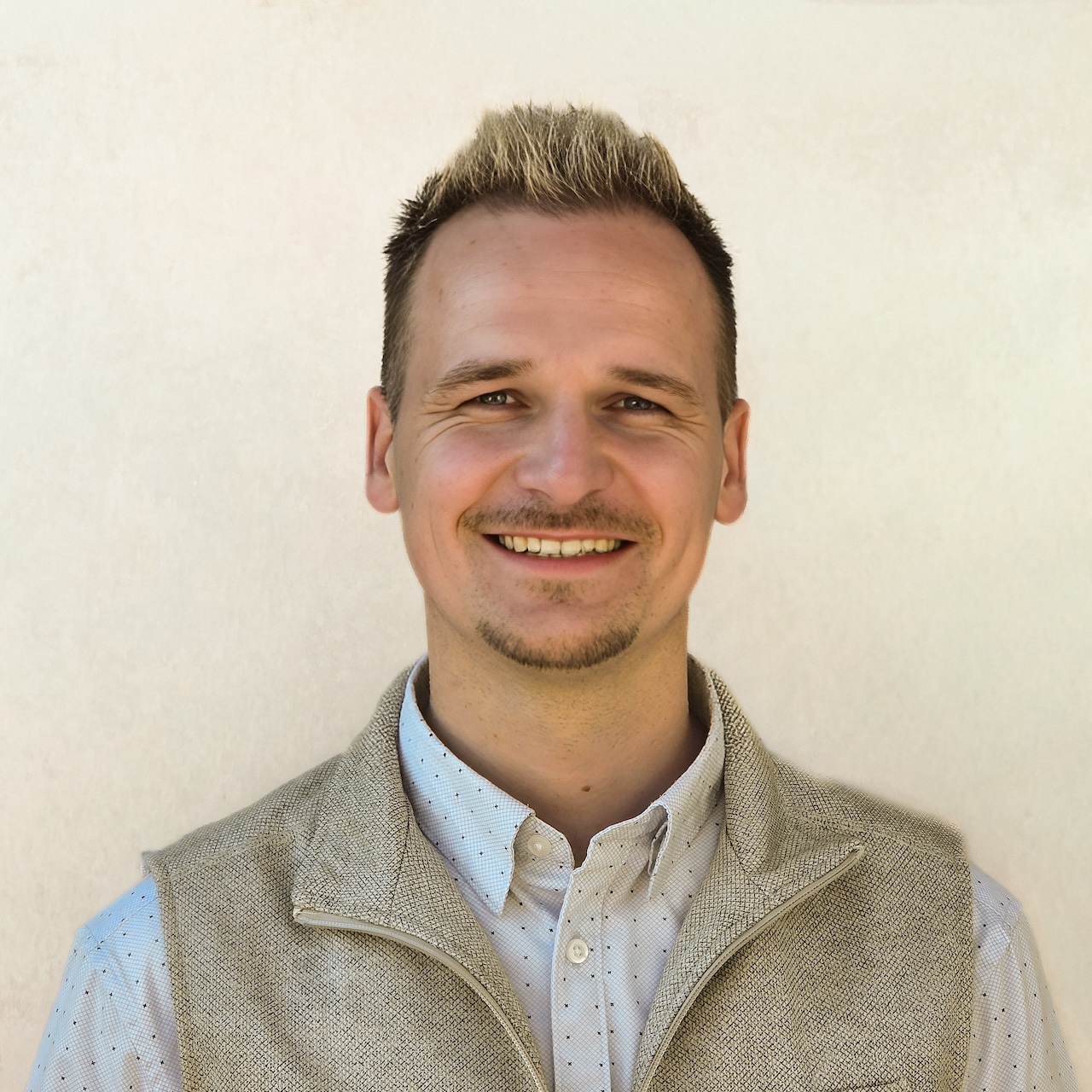 Smiling man in a light shirt and beige vest against a pale wall.