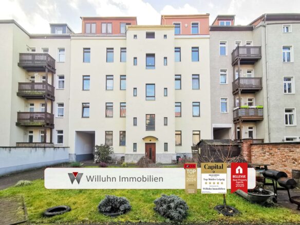 Front of a beige apartment building with balconies on both sides and a Willuhn Immobilien sign in the yard foreground.