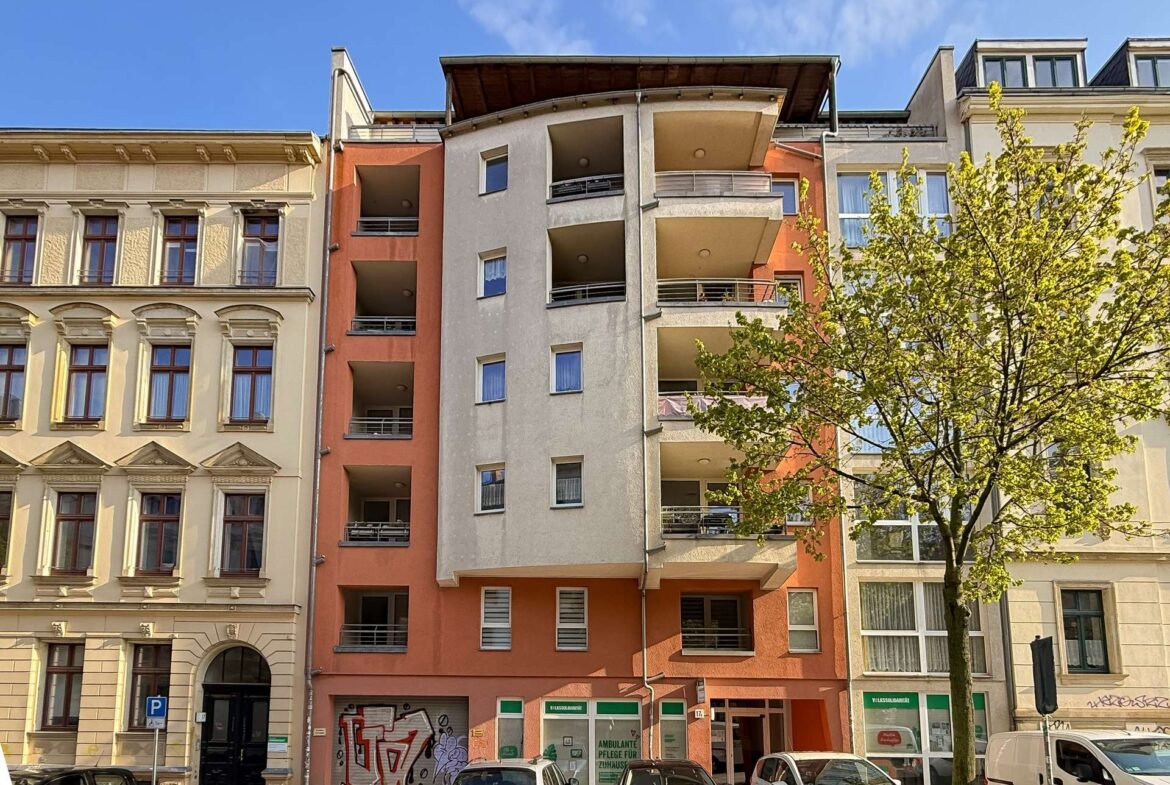 Street view of a colorful multi-story apartment building with balconies, parked cars, and a tree in front under a blue sky
