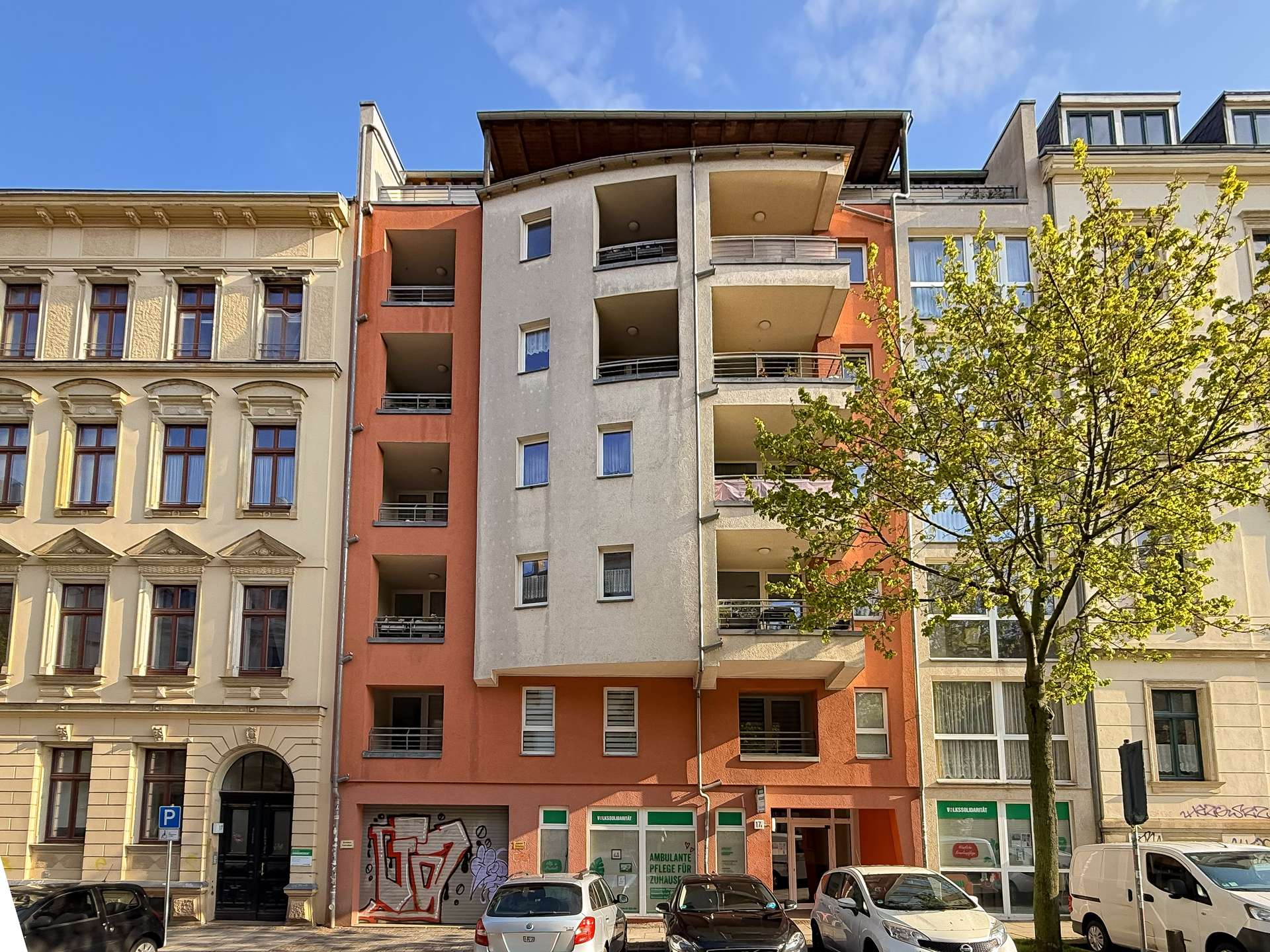 Street view of a colorful multi-story apartment building with balconies, parked cars, and a tree in front under a blue sky