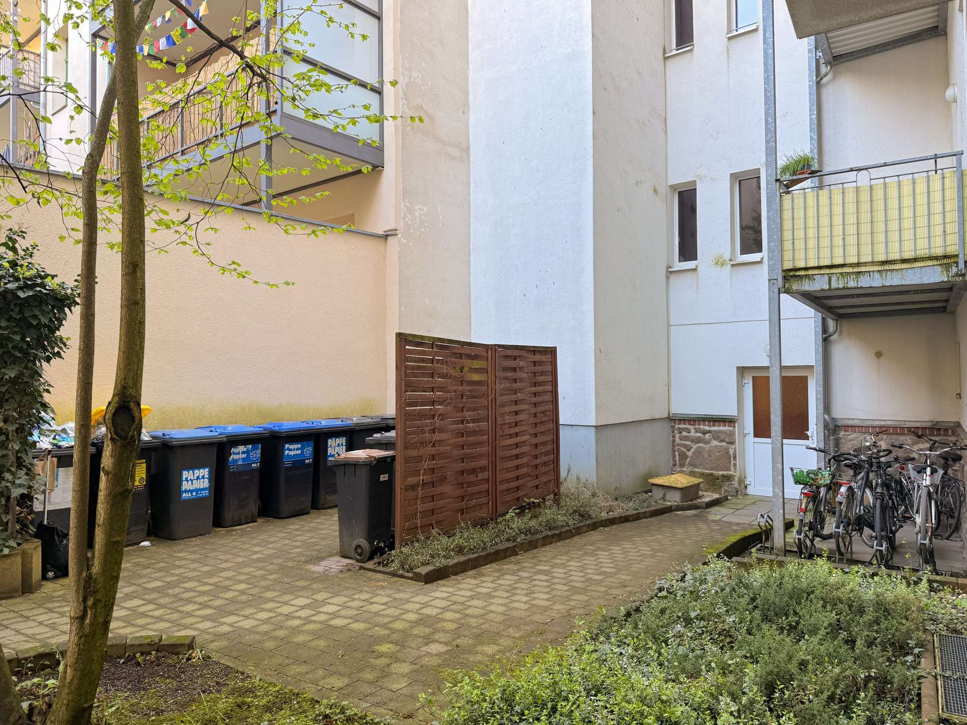 Urban courtyard between apartment buildings with blue recycling bins, a wooden fence, and several bicycles on the right, tree in foreground.
