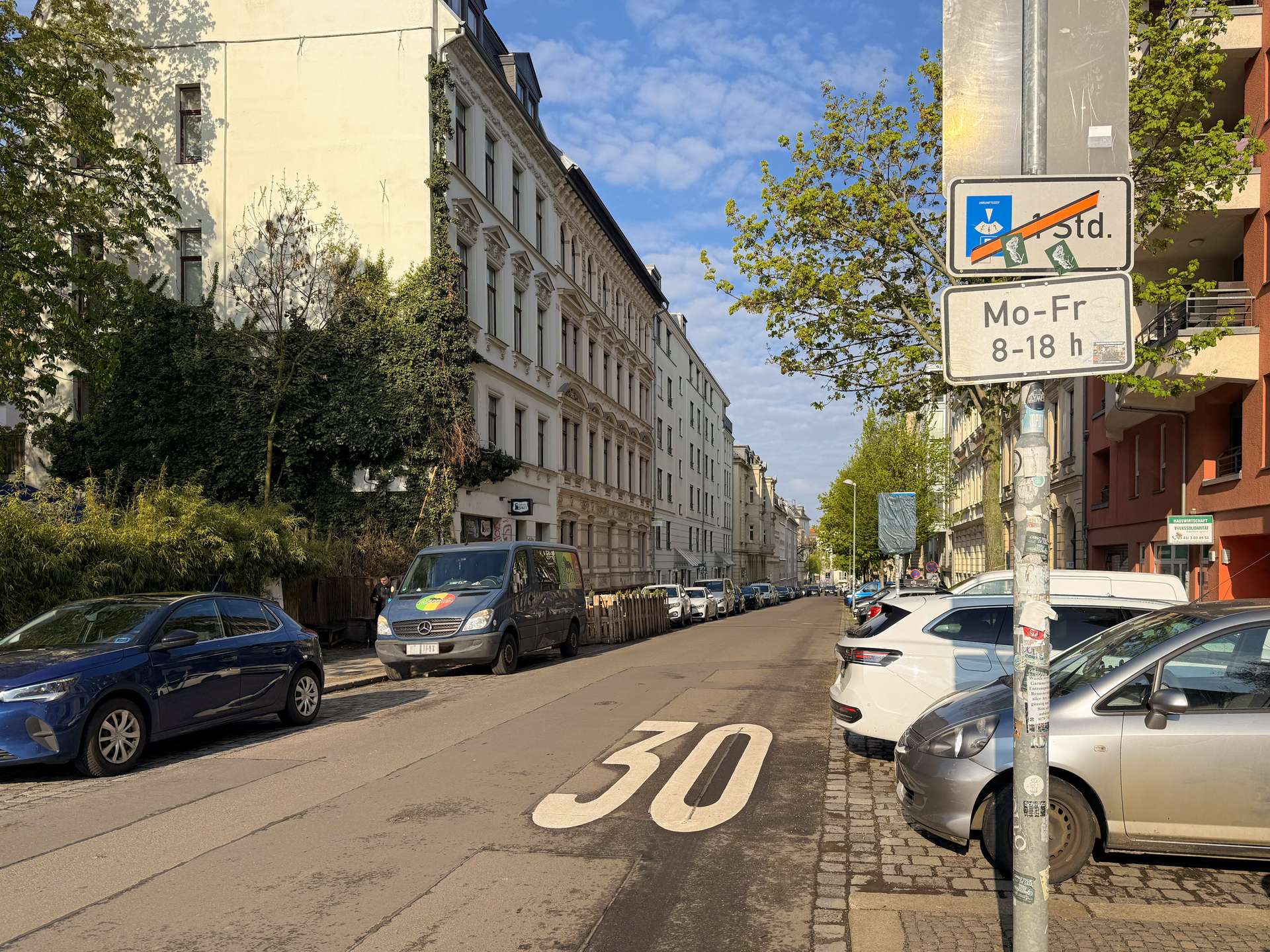 Urban street with parked cars, white multi-story buildings, and a large '30' speed limit marking on the road beneath a blue sky.