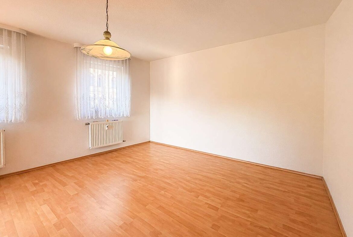 Empty room with light wood laminate flooring, white walls, two windows with sheer lace curtains, and a hanging ceiling light.