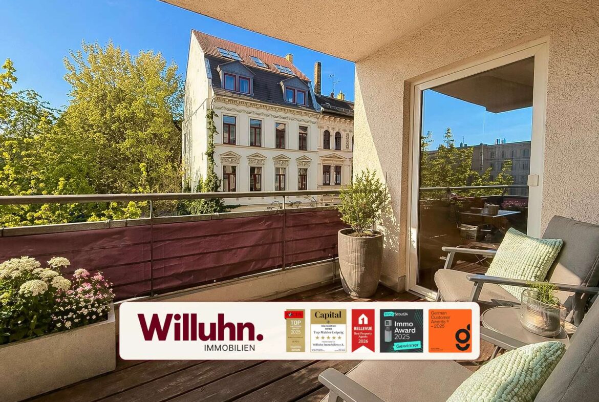 Sunny balcony with wooden deck, a beige wall, gray chair with green cushion, potted plants, and a view of a historic building across the street; Willuhn Immobilien banner at the bottom.