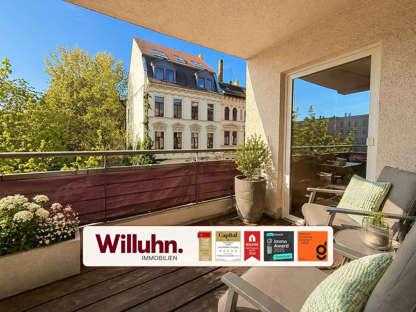 Sunny balcony with wooden deck, a beige wall, gray chair with green cushion, potted plants, and a view of a historic building across the street; Willuhn Immobilien banner at the bottom.