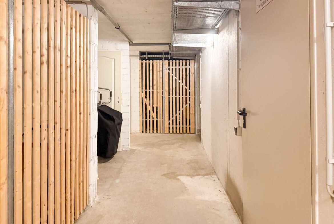 Concrete basement corridor with vertical wooden slat walls, a metal gate at the end, and a beige door on the right; exposed ceiling pipes.