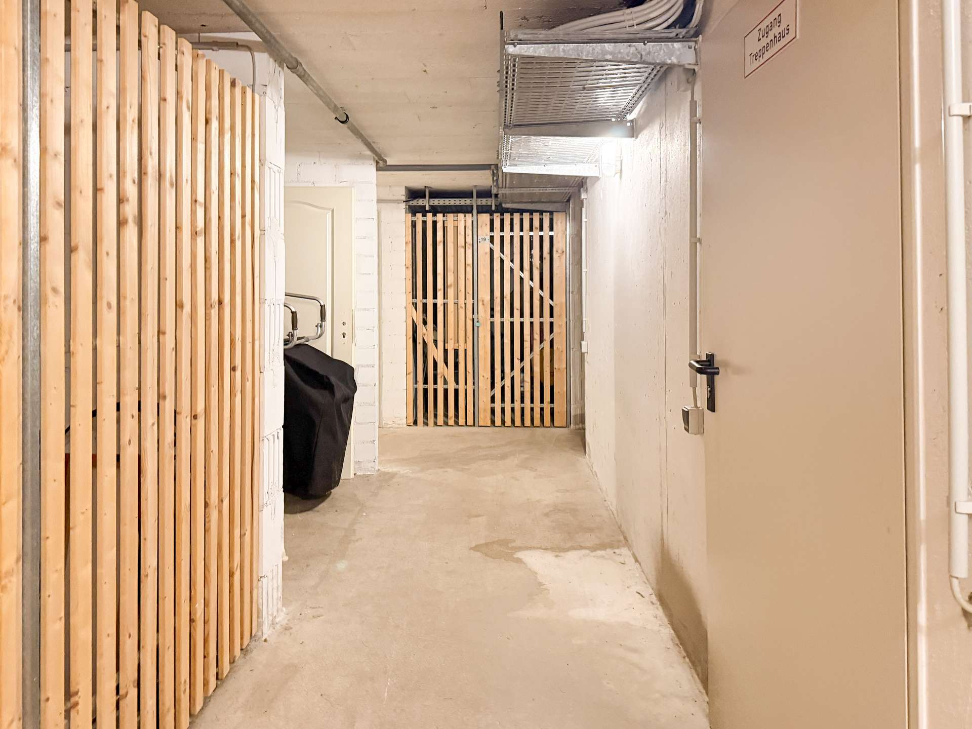 Concrete basement corridor with vertical wooden slat walls, a metal gate at the end, and a beige door on the right; exposed ceiling pipes.