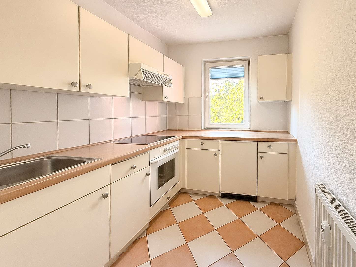 Compact L-shaped kitchen with white cabinets, beige countertops, and a window overlooking greenery