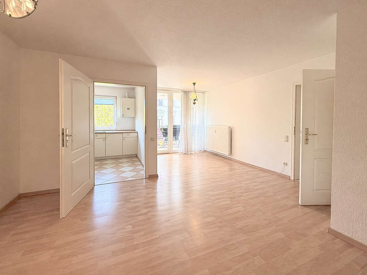 Bright, empty living room with light wood flooring, white walls, radiator, and a doorway to a small kitchen; glass doors to a balcony with sheer curtains in background.
