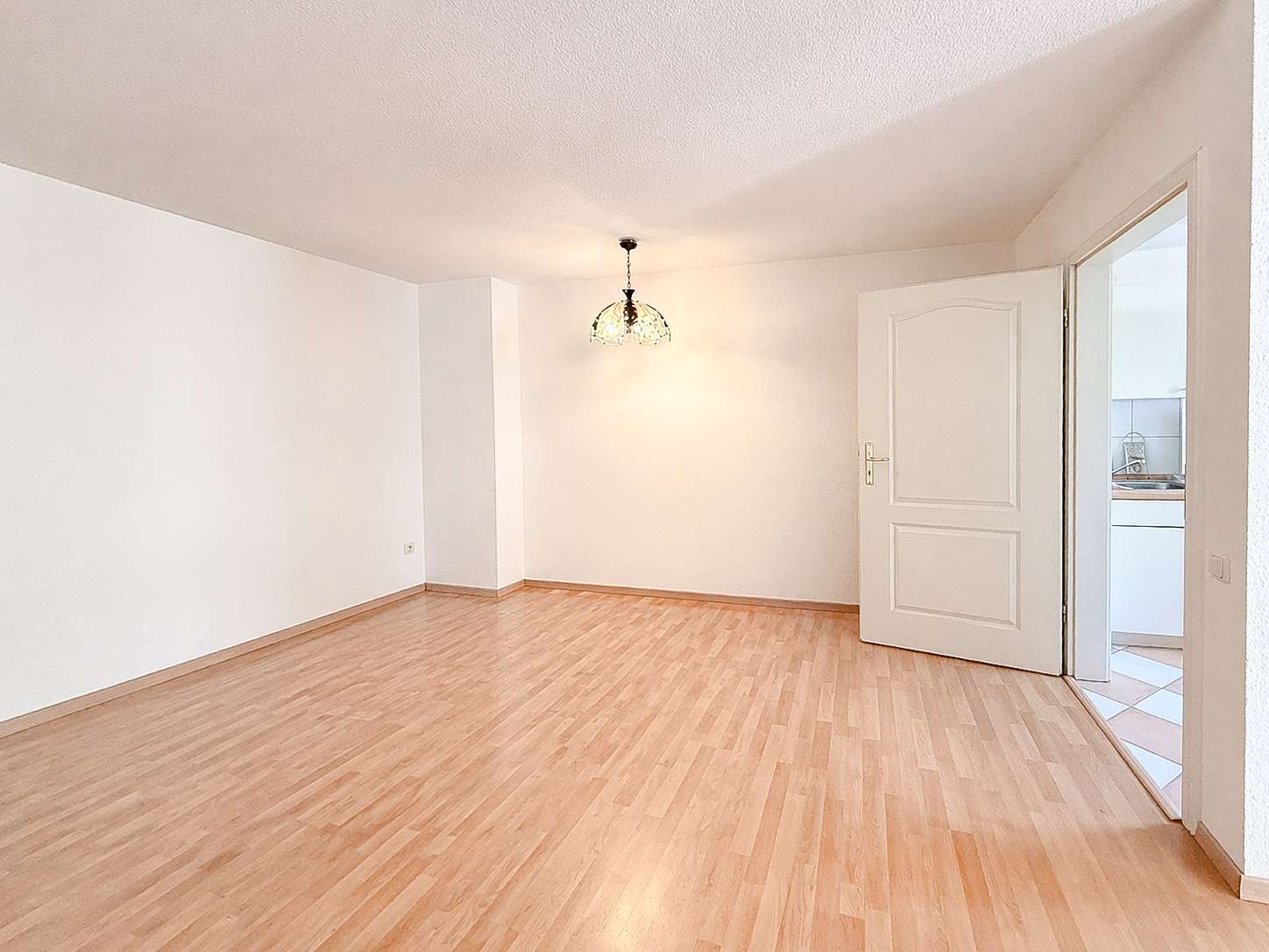 Empty living room with light wood flooring, white walls, a decorative ceiling light, and an open door to the kitchen.