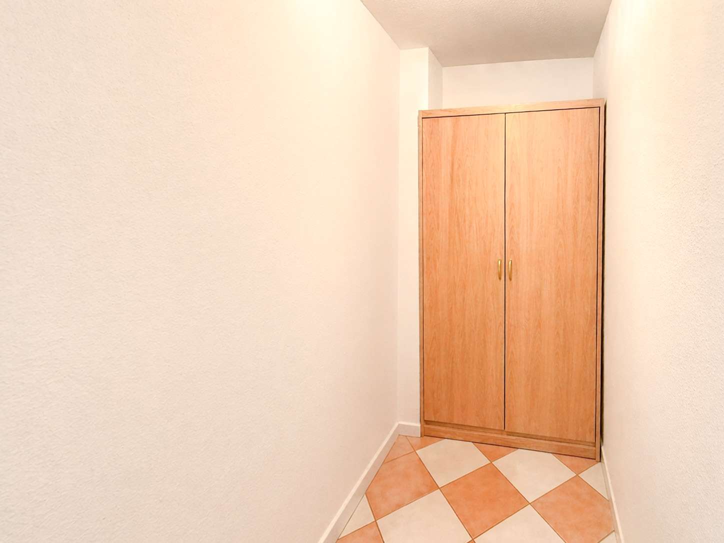 Narrow hallway with a tall wooden wardrobe at the end and a tiled orange-and-cream floor, walls painted white.