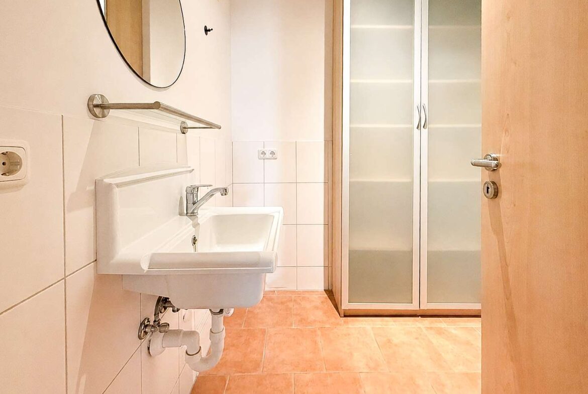 Compact bathroom with a wall-mounted white sink, circular mirror, towel bar, and exposed plumbing under a tiled wall. A frosted-glass cabinet is visible at the far end with a wooden door nearby.