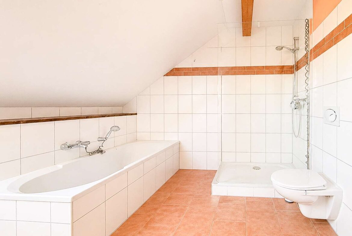 Bright bathroom with a white tiled bathtub along the left wall under a sloped ceiling; terracotta floor tiles and a separate shower area with white and orange tiles on the far wall; wall-mounted toilet on the right.