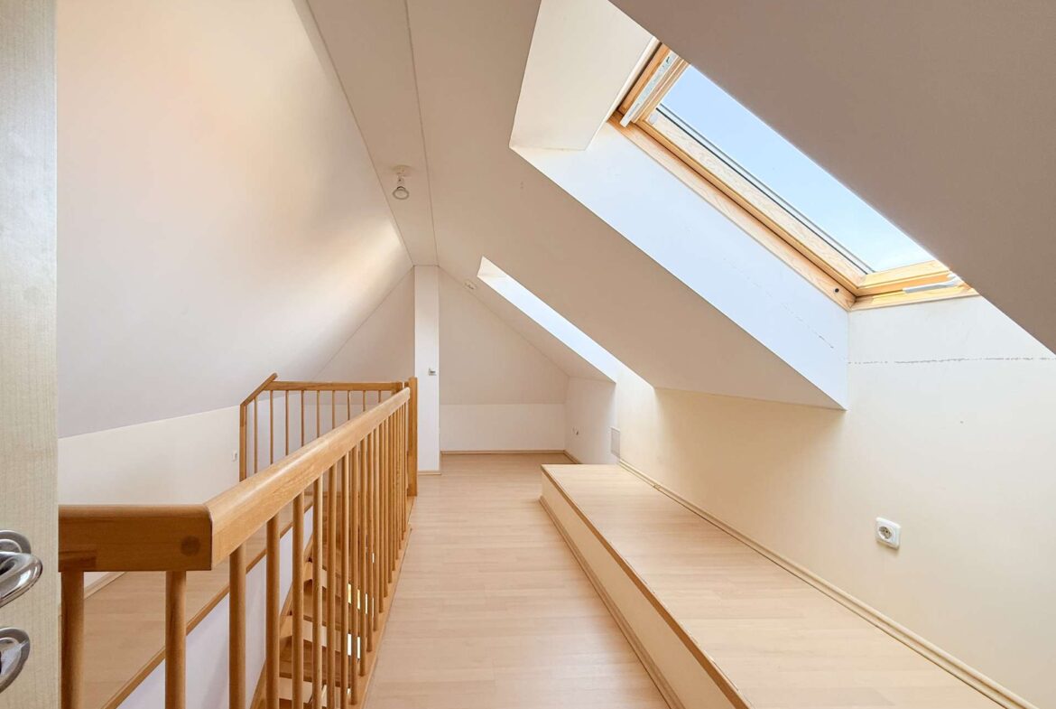 Bright attic corridor with sloped white walls, a wooden railing on the left, and a large skylight.