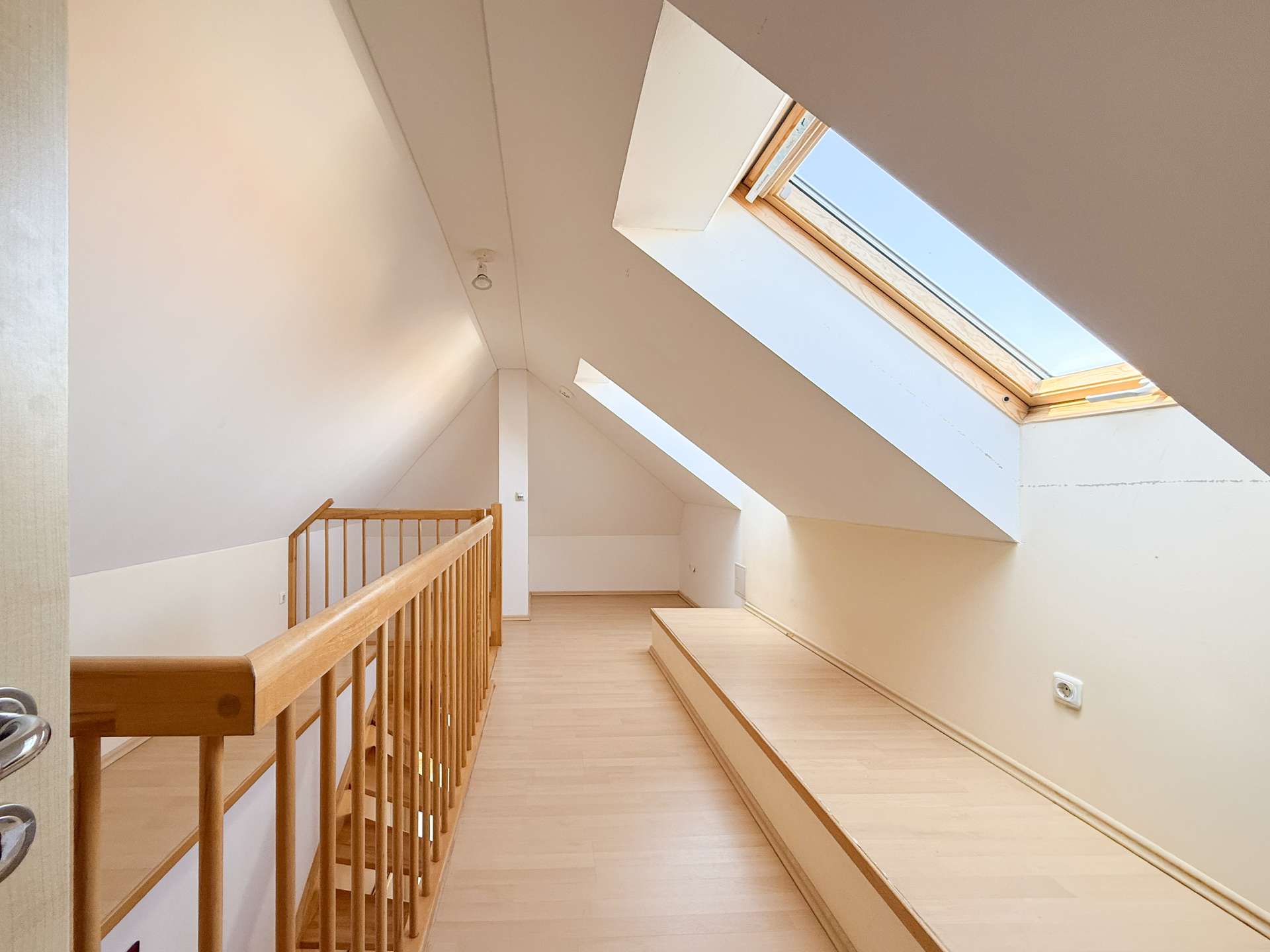 Bright attic corridor with sloped white walls, a wooden railing on the left, and a large skylight.