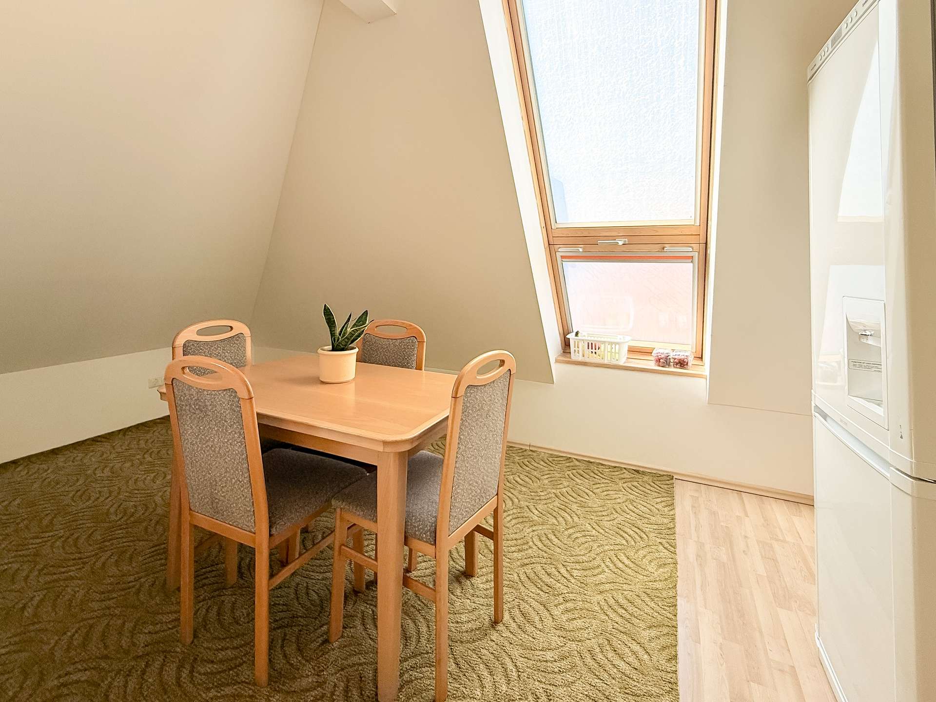 Dining area under a slanted ceiling with a light wood table, four upholstered chairs, and a potted plant on the table beneath a large skylight window in natural light.