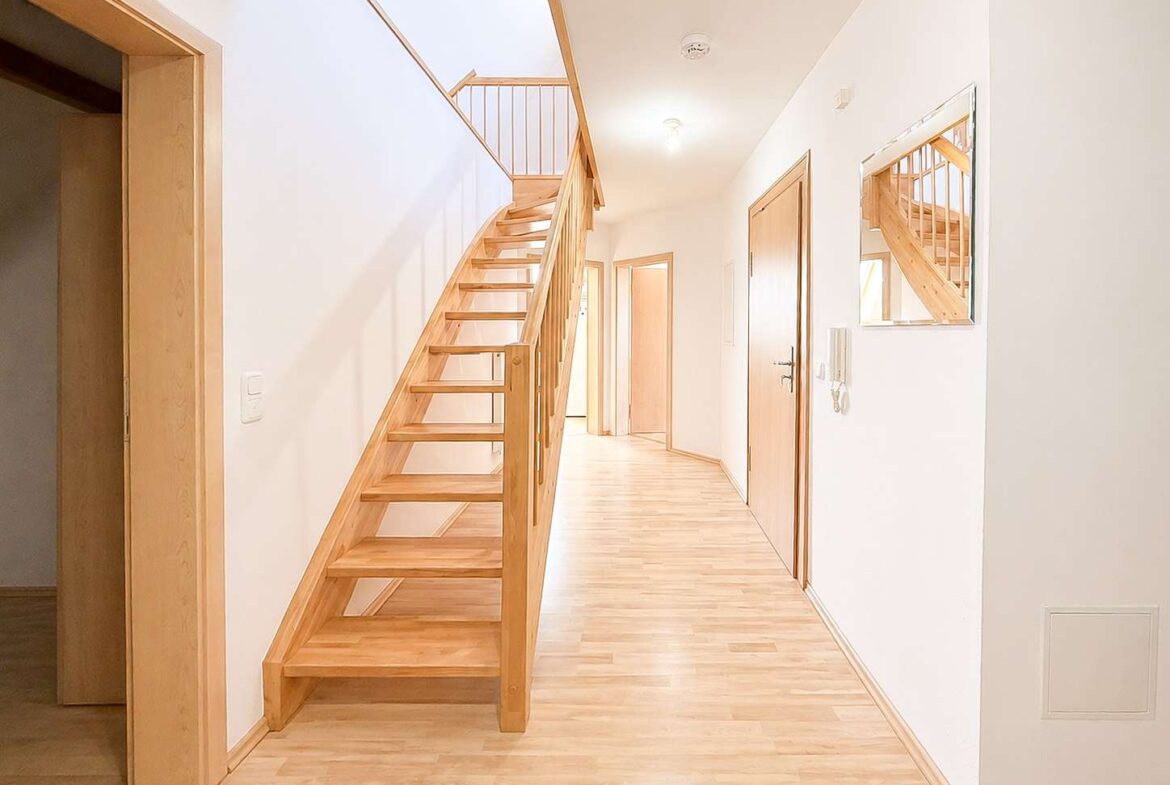 Bright hallway with a wooden staircase on the left and several closed wooden doors along the right wall, mirrored by a large wall mirror.