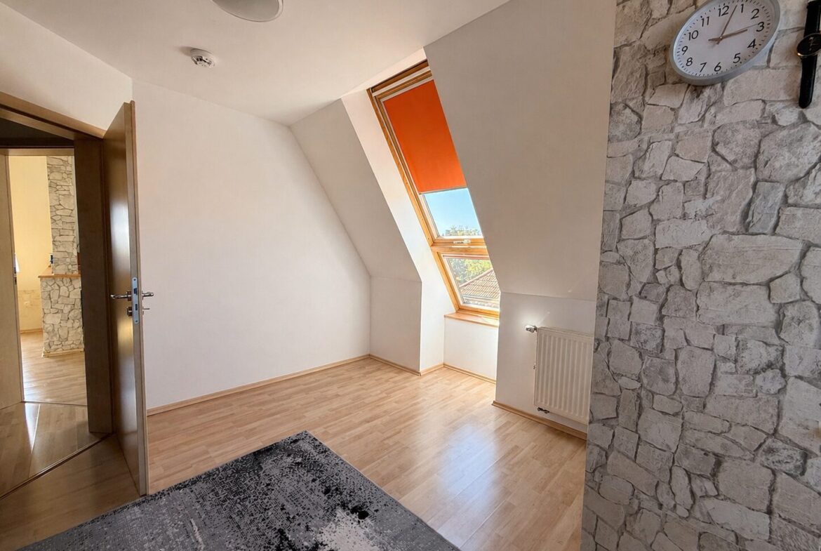 Bright attic room with a slanted ceiling, large skylight window with orange shade, and light wood flooring.