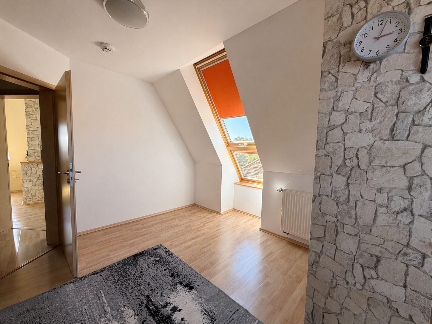 Bright attic room with a slanted ceiling, large skylight window with orange shade, and light wood flooring.
