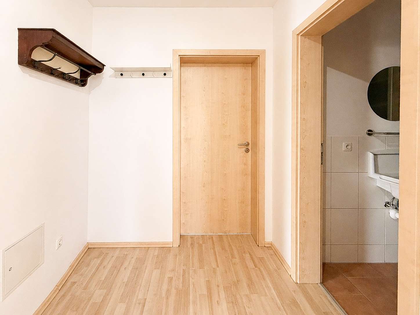 Hallway with light wood floor, two wooden doors, left wall coat rack, and open bathroom door on the right.