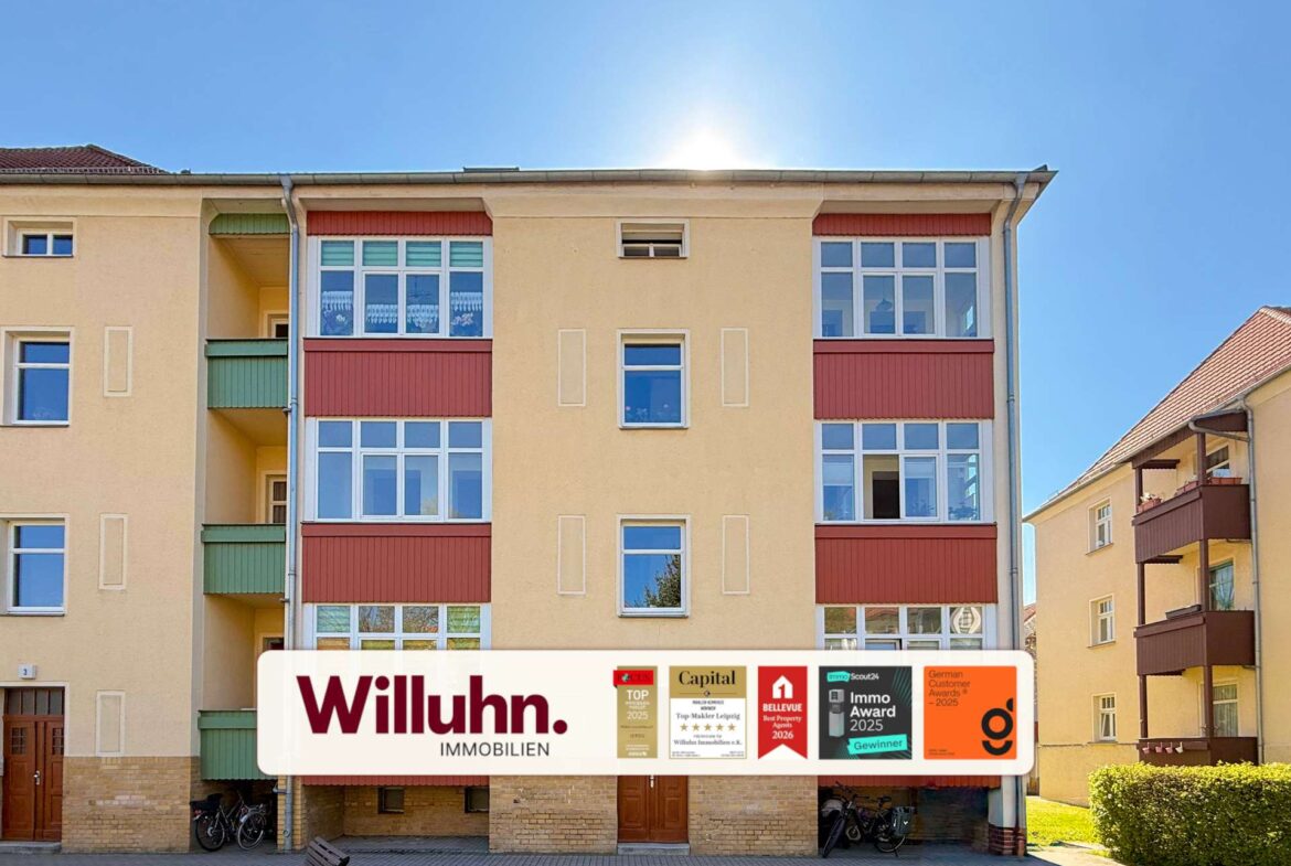 Beige multi-story apartment building with a large Willuhn Immobilien real estate banner and award logos on the ground floor storefronts under a blue sky.