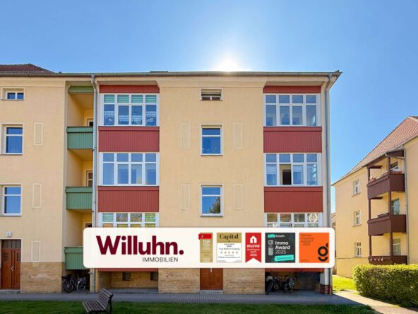 Beige multi-story apartment building with a large Willuhn Immobilien real estate banner and award logos on the ground floor storefronts under a blue sky.