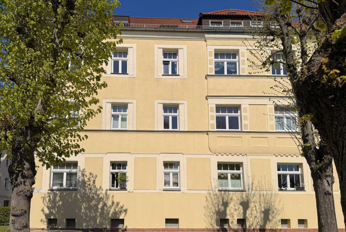 Facade of a pale yellow multi-story building with white-framed windows, flanked by green trees and a blue sky above.